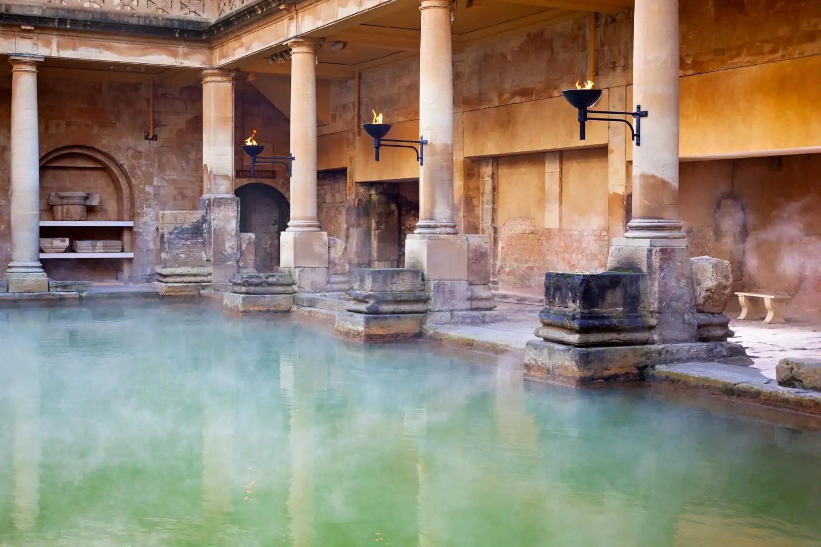Great Bath v Roman Baths s antickými sloupy, hořícími pochodněmi a párou nad zelenavou termální vodou